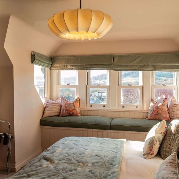 Gemütliches Schlafzimmer mit einer Fensternische, grünen Kissen, einem Bett mit gemusterten Kissen, einer sanften Lampe und einem hängenden Bademantel. Große Fenster mit Blick auf die Hügel draußen.