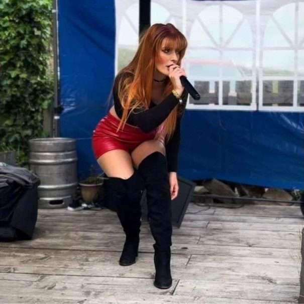 Frau mit langen roten Haaren, die ein schwarzes Oberteil, einen roten Rock und schwarze kniehohe Stiefel trägt, singt auf einer Freilichtbühne in ein Mikrofon.