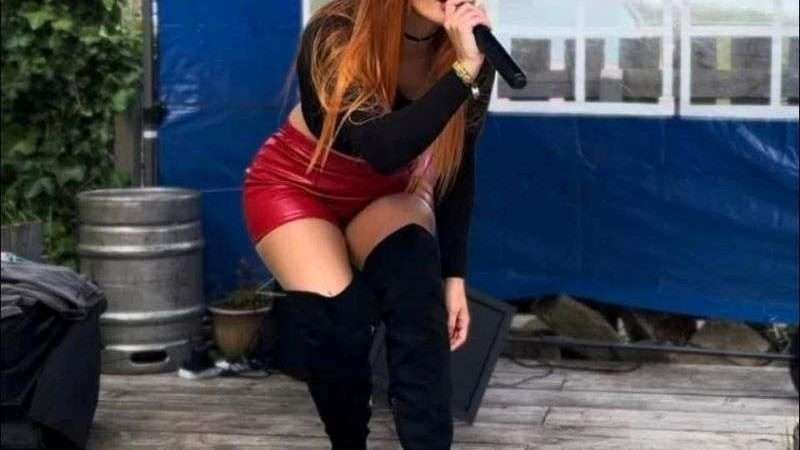 Frau mit langen roten Haaren, die ein schwarzes Oberteil, einen roten Rock und schwarze kniehohe Stiefel trägt, singt auf einer Freilichtbühne in ein Mikrofon.