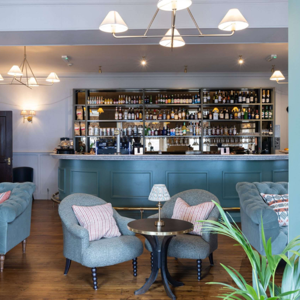 Un bar lounge avec des canapés et fauteuils bleu sarcelle, une table ronde avec une petite lampe, un parquet et un bar bien garni en arrière-plan.