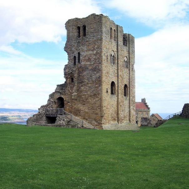 Un donjon de château en pierre partiellement ruiné se dresse sur une colline herbeuse avec quelques bâtiments à l'arrière-plan sous un ciel partiellement nuageux.