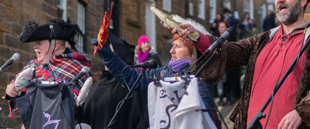 Drei verkleidete Personen singen und gestikulieren auf einer Open-Air-Bühne mit Mikrofonen; Banner hängen auf Ständern vor ihnen, und alte Steingebäude säumen den Hintergrund.