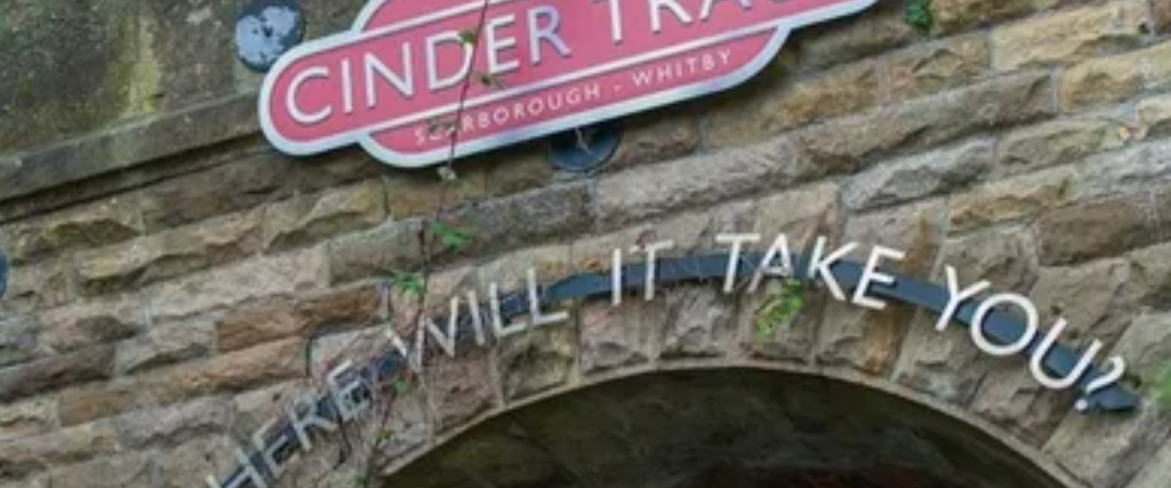 Stone archway with a sign reading "The Cinder Track Scarborough Whitby" and the phrase "Where will it take you?" below it, surrounded by greenery.