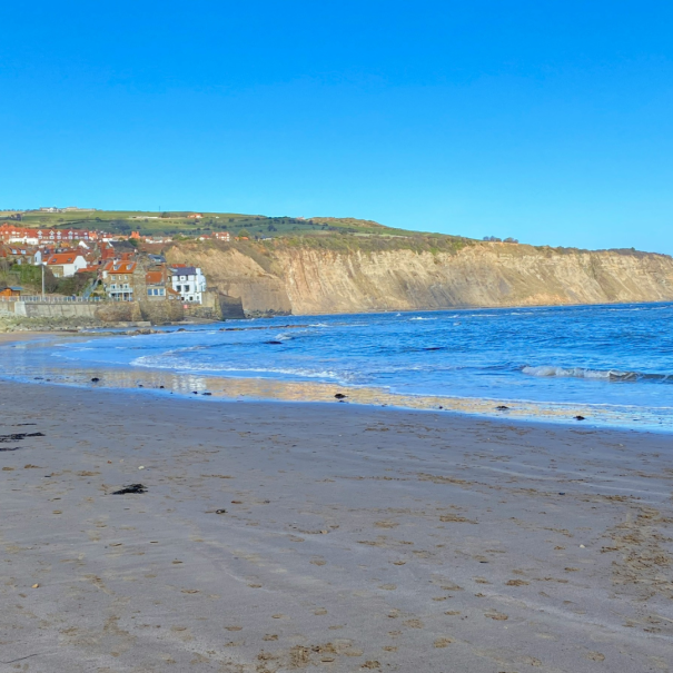 A sandy beach with gentle waves, cliffs in the background, and houses visible on the grassy hill under a clear blue sky.