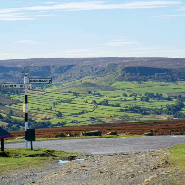 Eine Landstraße mit einem Wegweiser nach Lealholm und Guisborough, mit Blick auf grüne, flickenteppichartige Felder und ferne Hügel unter einem klaren Himmel.