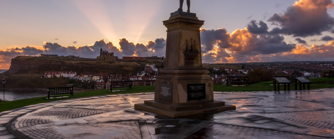 Eine Statue steht bei Sonnenaufgang auf einer runden Steinplattform, mit Sonnenstrahlen und Wolken am Himmel und einer Stadt in der Ferne.