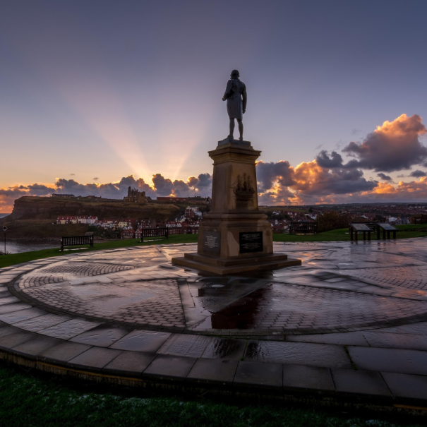 Eine Statue steht bei Sonnenaufgang auf einer runden Steinplattform, mit Sonnenstrahlen und Wolken am Himmel und einer Stadt in der Ferne.