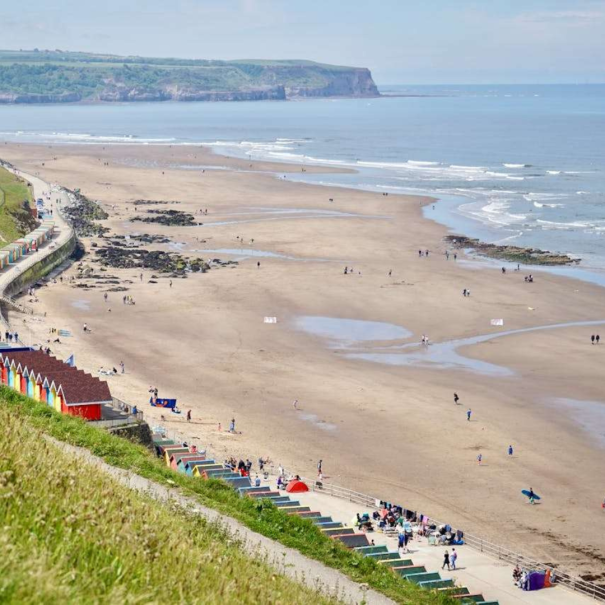 A wide sandy beach with scattered people, colorful beach huts along the promenade, and cliffs in the distance under a partly cloudy sky.