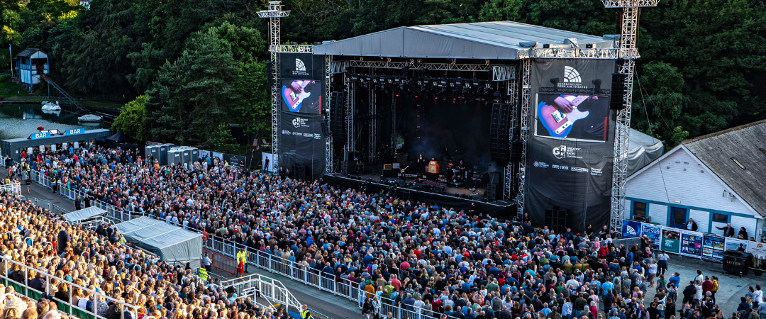 Eine große Menschenmenge verfolgt ein Live-Musikkonzert auf einer Open-Air-Bühne, umgeben von Bäumen unter einem teilweise bewölkten Himmel.