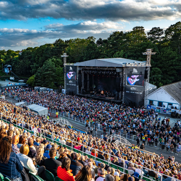Eine große Menschenmenge verfolgt ein Live-Musikkonzert auf einer Open-Air-Bühne, umgeben von Bäumen unter einem teilweise bewölkten Himmel.