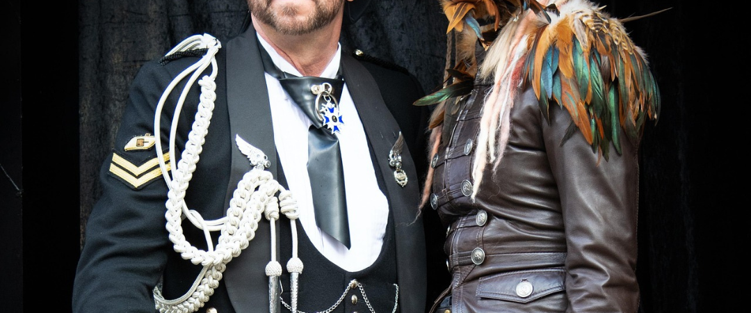 Zwei Personen in Steampunk-inspirierten Outfits mit dekorativen Accessoires und Sonnenbrillen, die vor einem dunklen Hintergrund zusammenstehen.