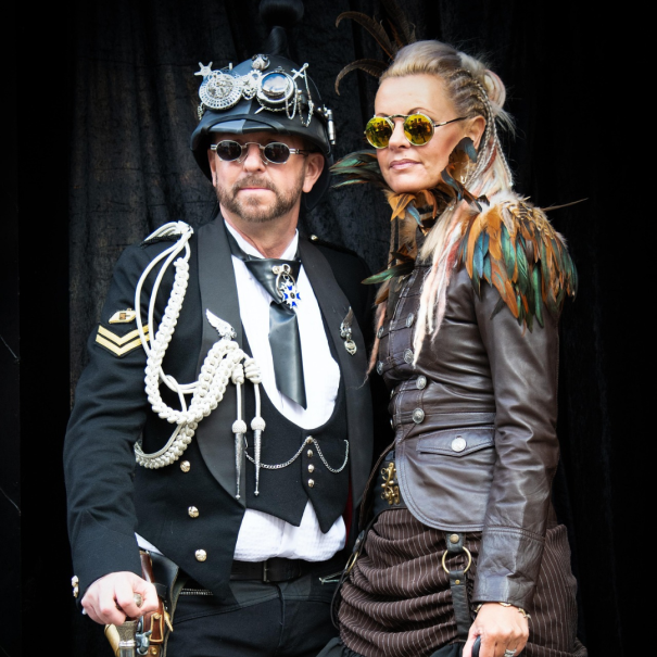 Zwei Personen in Steampunk-inspirierten Outfits mit dekorativen Accessoires und Sonnenbrillen, die vor einem dunklen Hintergrund zusammenstehen.