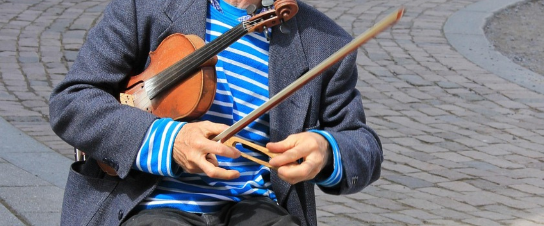 Ein Mann mit einer blauen Mütze und einem gestreiften Hemd sitzt draußen auf einem Stuhl und hält eine Violine und einen Bogen, während er scheinbar auf einer kopfsteingepflasterten Straße Musik spielt.