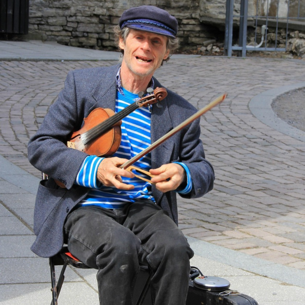 Ein Mann mit einer blauen Mütze und einem gestreiften Hemd sitzt draußen auf einem Stuhl und hält eine Violine und einen Bogen, während er scheinbar auf einer kopfsteingepflasterten Straße Musik spielt.