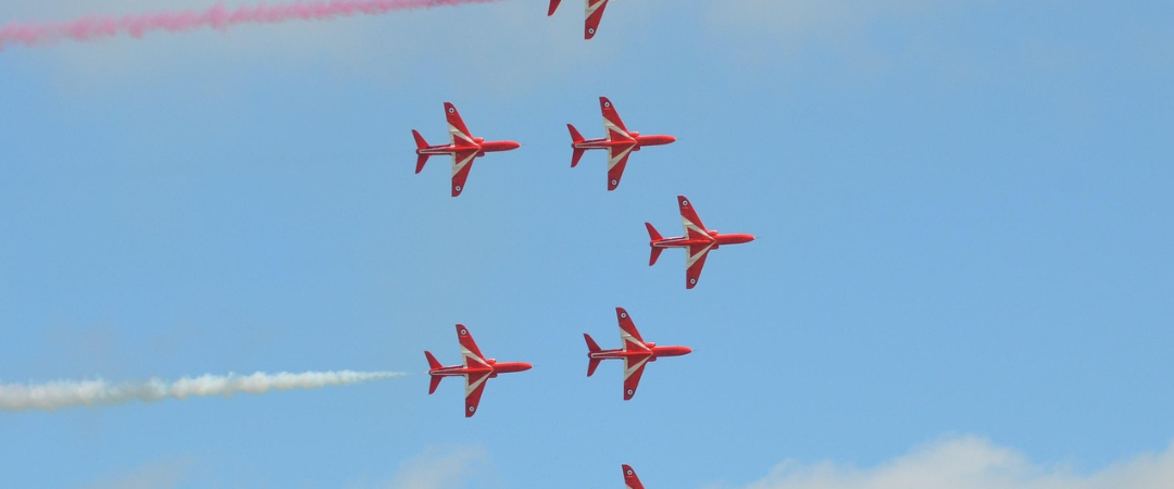 Sieben rote Jets fliegen in Formation und stoßen farbige Rauchschwaden aus – rot, weiß und blau – vor einem blauen Himmel mit vereinzelten Wolken.