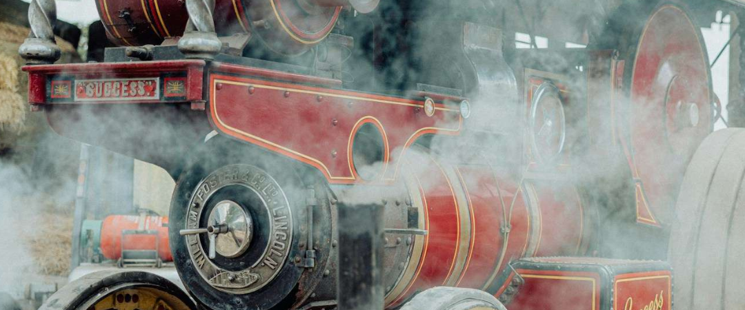 A vintage steam-powered traction engine with red and gold detailing stands amid clouds of steam, displaying the name "L.C. Byass & Son" and "Success" on its side.